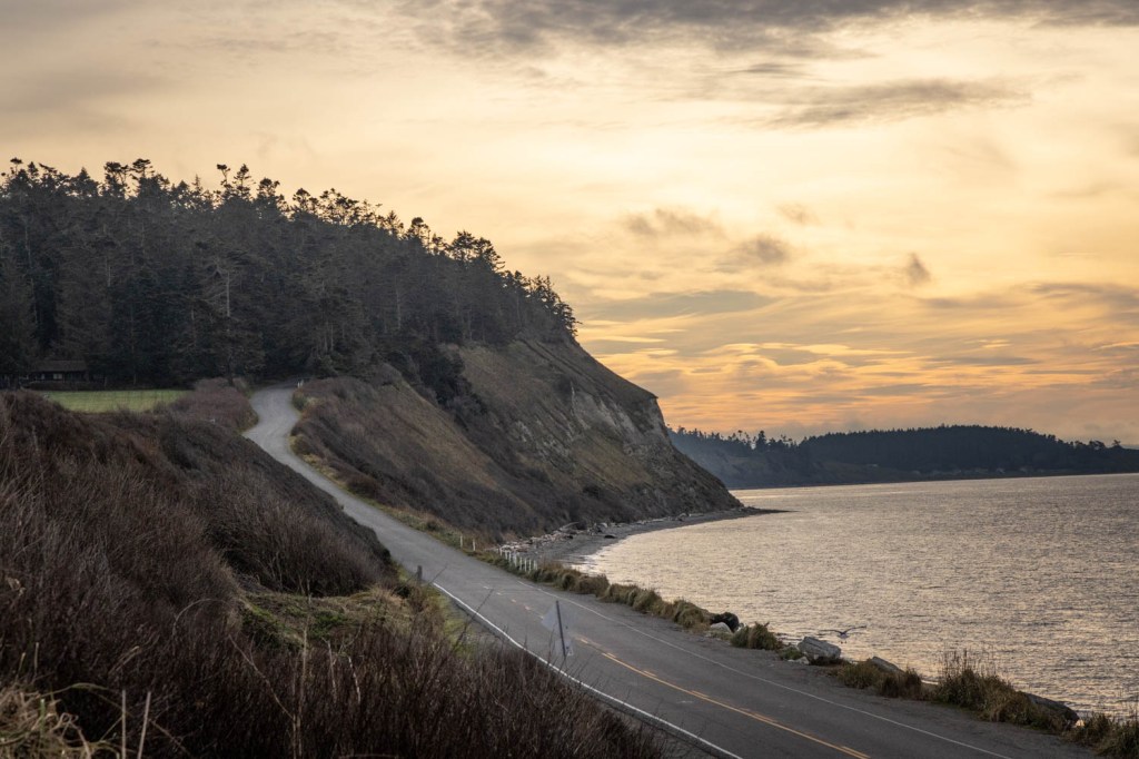A winding road along a coastal cliff with forested land and a calm sea under a cloudy sunset sky.